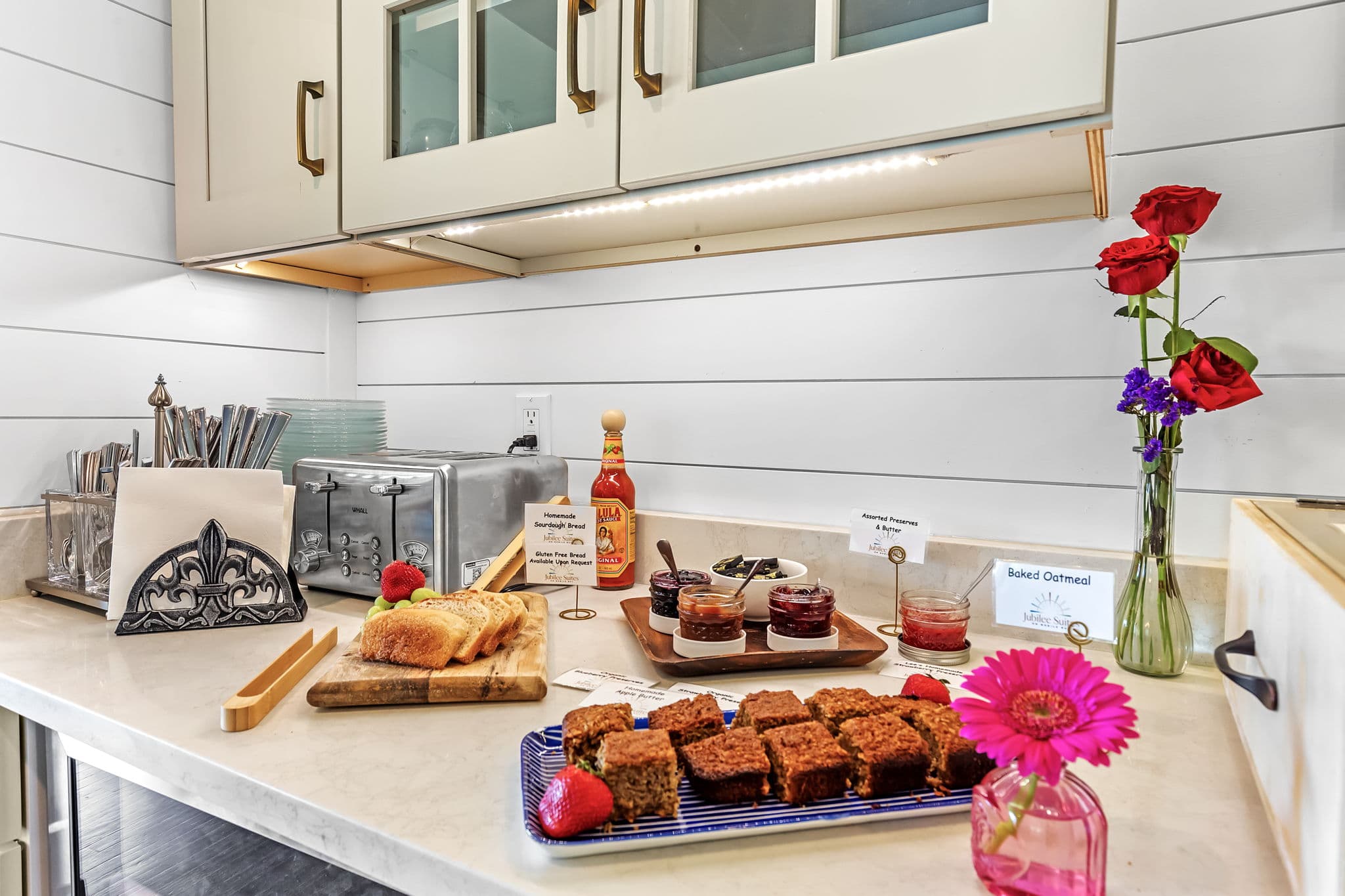 A kitchen counter with a spread of baked goods and other treats. There are plates of brownies, a loaf of bread, and a jar of jam. Two vases of flowers and a hot sauce bottle add a touch of color.