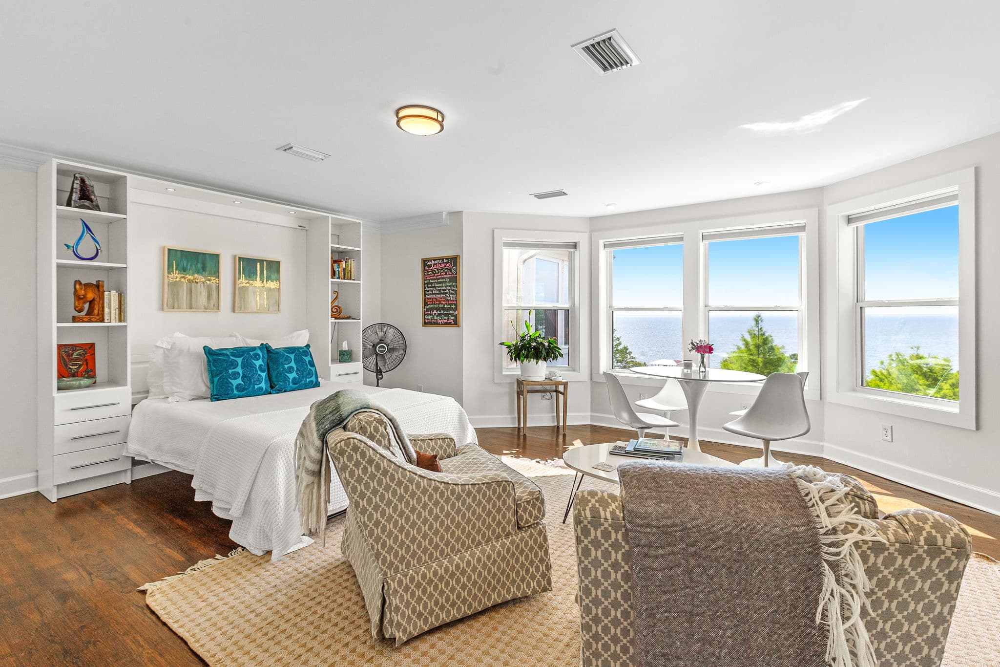 A spacious living area with large bay windows overlooking the ocean. The room features a comfortable murphy bed, seating area, and built-in bookshelves.