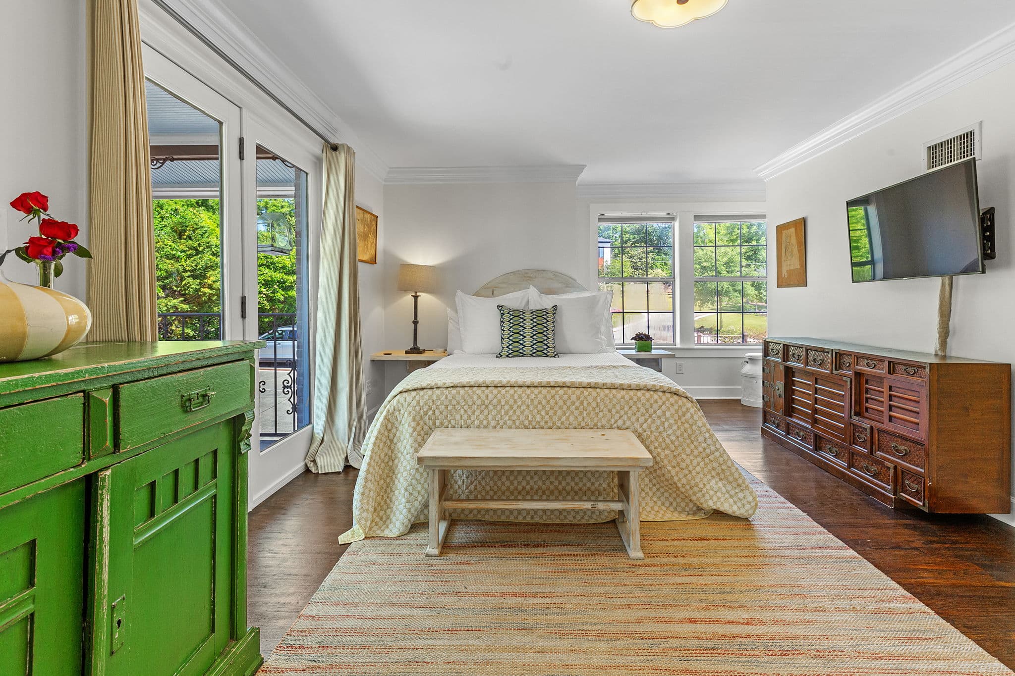 A spacious bedroom with sliding glass doors leading to a lush garden. The room features a comfortable bed, a green dresser, and a wooden bench.