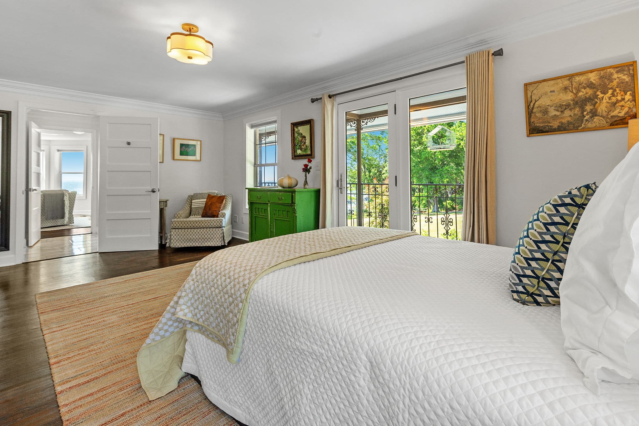 A spacious bedroom with large windows overlooking a lush garden. The room features a comfortable bed, a green dresser, and a wooden bench.