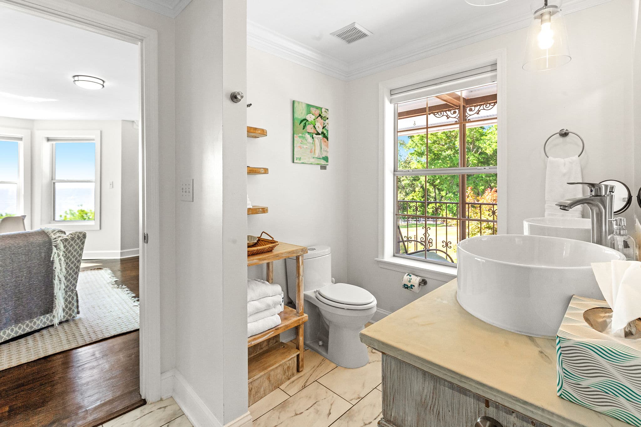 A spacious bathroom with large windows overlooking the bay. The room features a white sink, a toilet, and a wooden shelf with towels.
