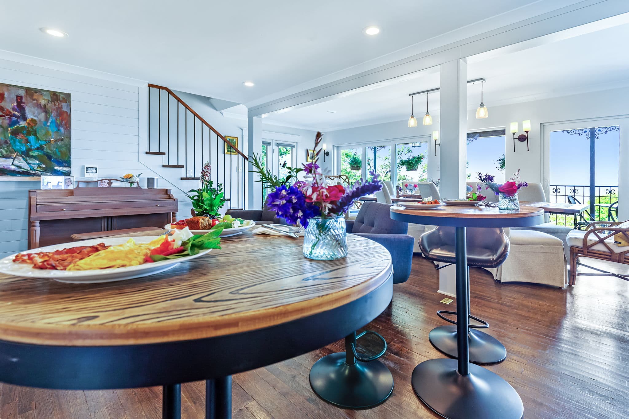 A spacious living room featuring a breakfast buffet with various dishes, a piano, a staircase, and a view of a balcony with outdoor seating and a garden