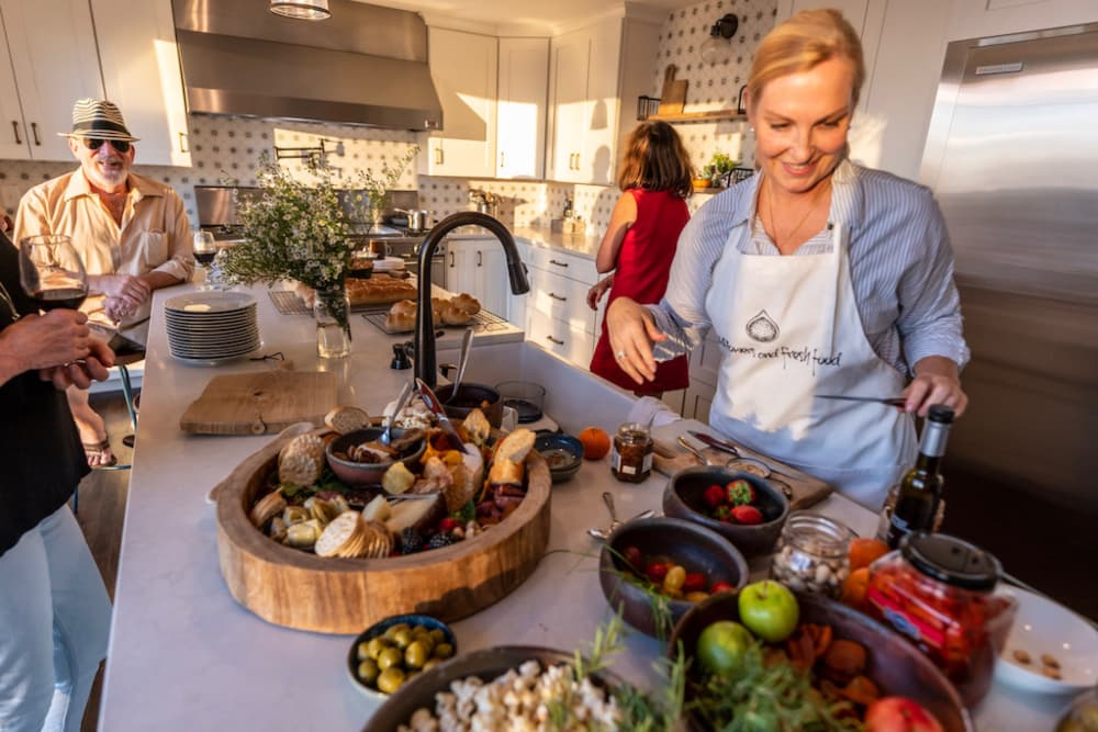 A large kitchen with a woman arranging beautiful foods for a catered meal, with other people standing around ready to fill their plates.