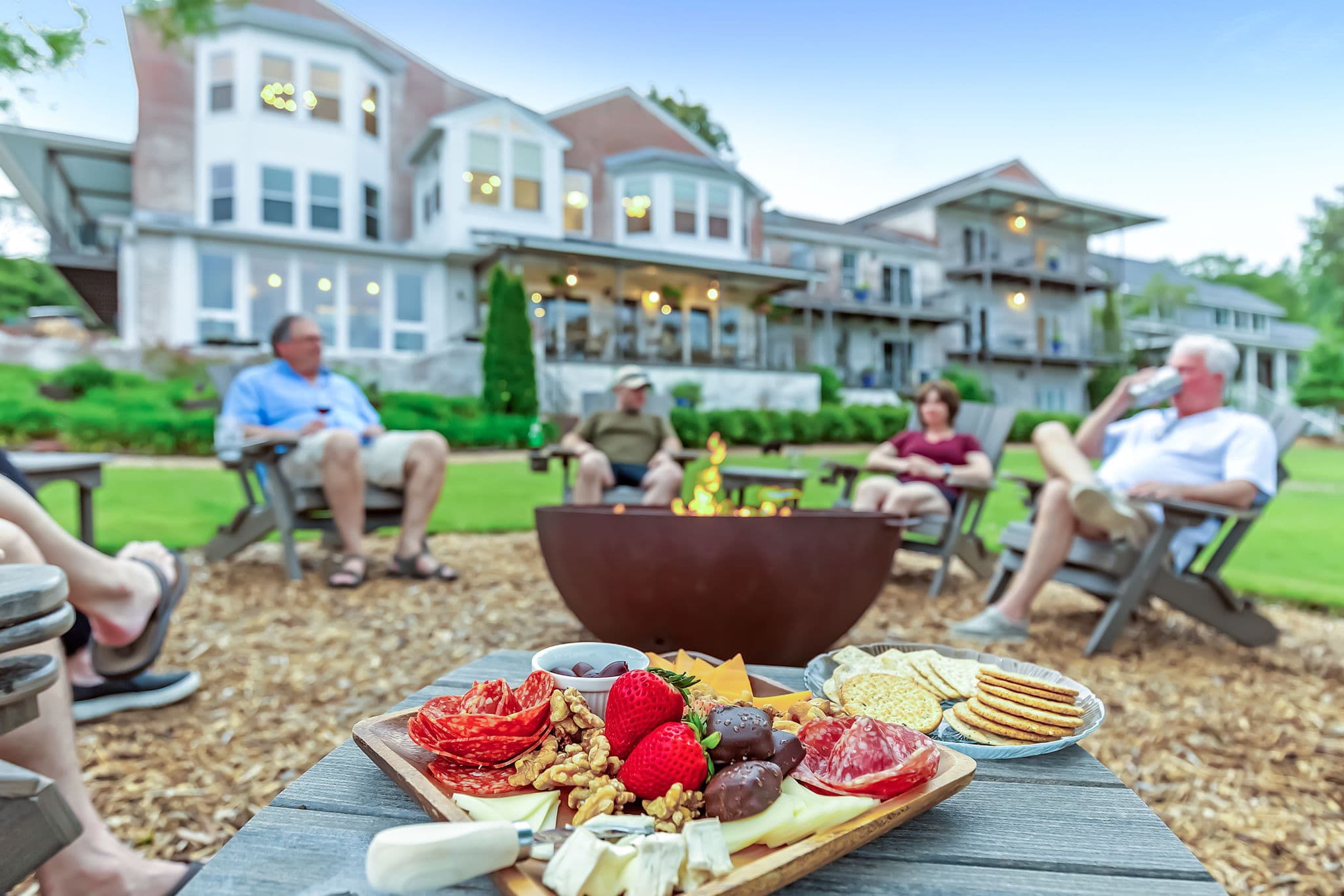 A cozy outdoor gathering with friends around a fire pit, featuring a charcuterie board in the foreground and a large house in the background.