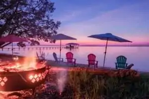 A serene lakeside scene at sunset with a fire pit, colorful chairs, and umbrellas.