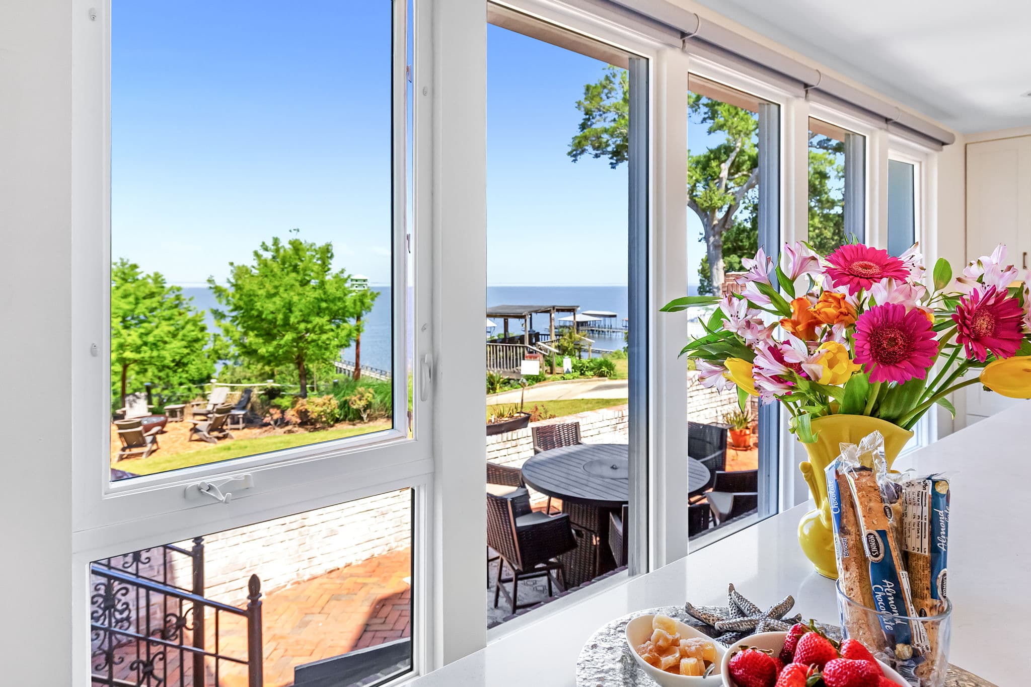 Large windows in a kitchen offering a view of a calm body of water, a patio with outdoor seating, and a grassy area with trees.
