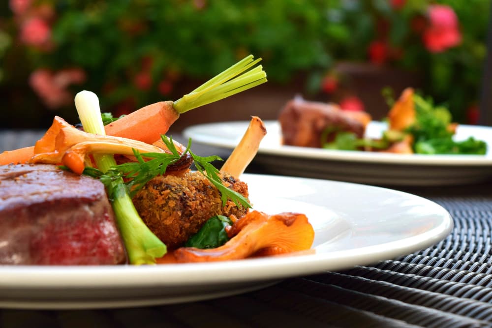 Two plates with elegantly plated steaks, racks of lamb, and colorful vegetables.