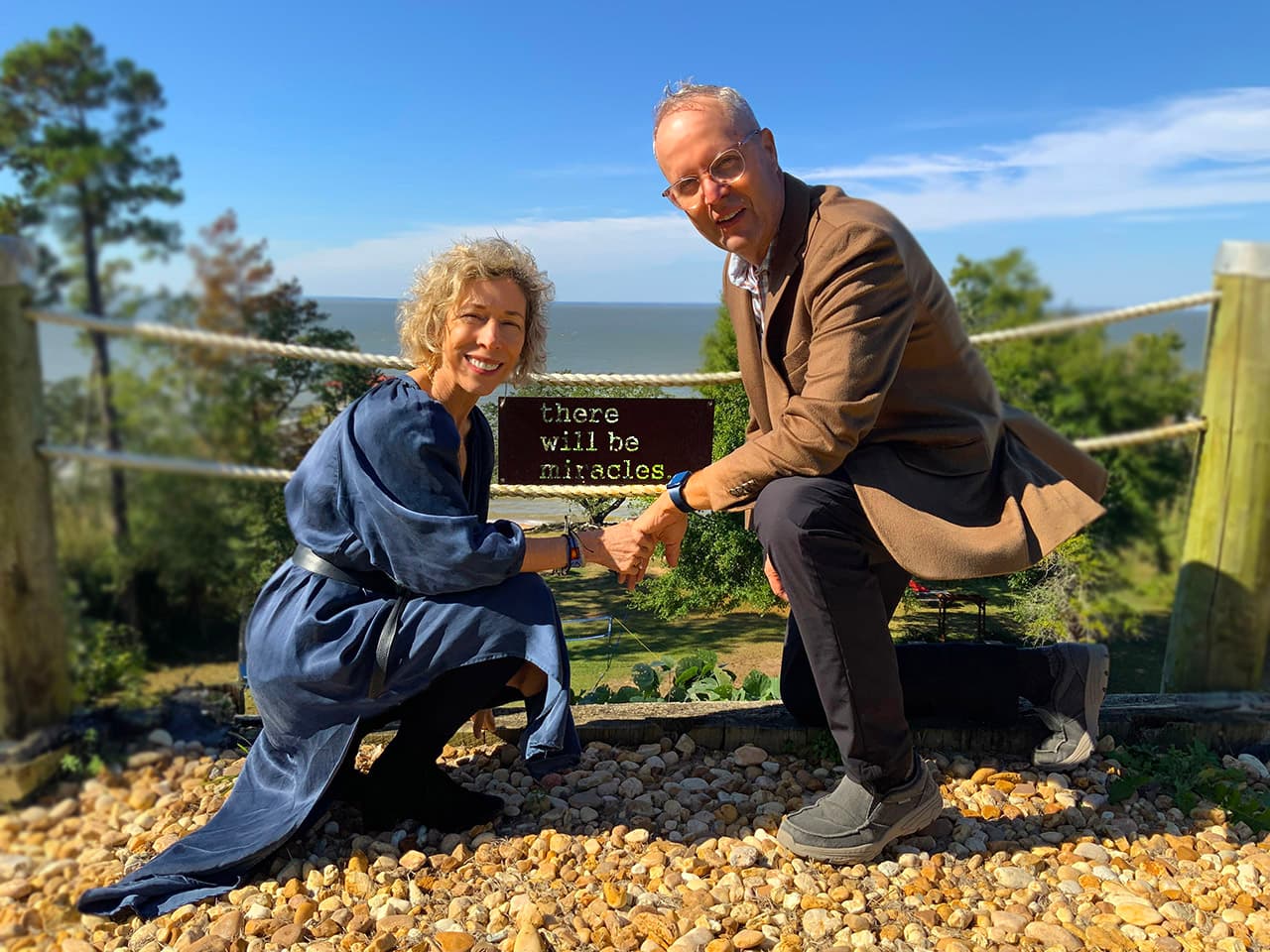 A woman and a man kneel together outdoors, smiling as they hold a sign that reads "there will be miracles."