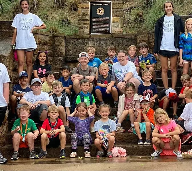 A large group of children and adults, some wet, poses together on stone steps near a plaque. A large group of children and adults, some wet, poses together on stone steps near a plaque.