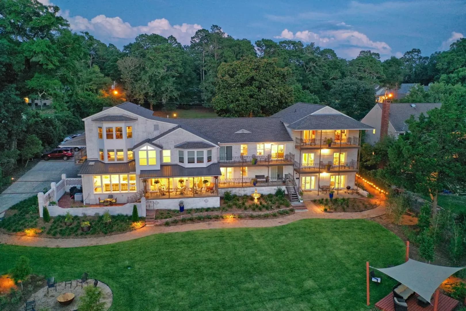 Aerial view of a large, illuminated house surrounded by greenery at dusk.