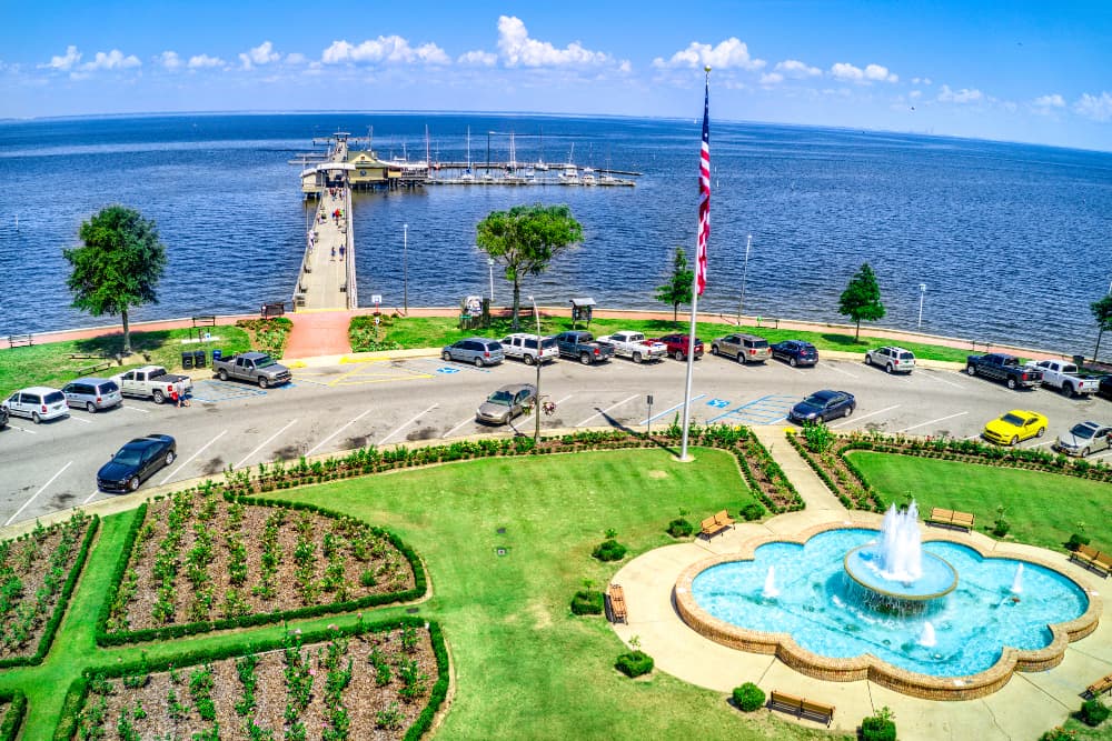 A beautiful view of the blue waters of Mobile Bay with a pier and dock with people walking on the pier, and a green grassy area with trees and an American flag in the foreground.