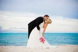 A groom kisses a bride in a white wedding dress with a bouquet at the beach.