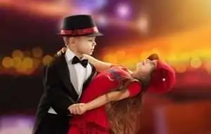 A boy in a tuxedo dances with a girl wearing a red dress and hat against a colorful background.