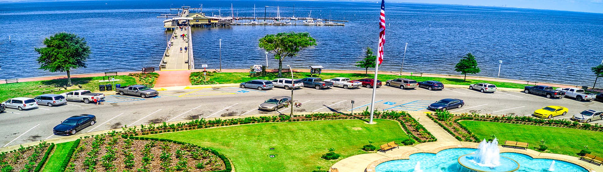 A waterfront view featuring a pier, parked cars, and a landscaped area with a fountain.
