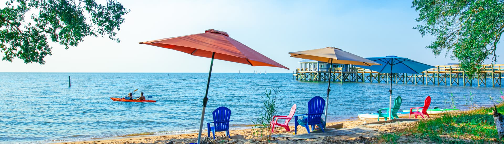 A grassy area that turns to a sandy beach on the shores of Mobile Bay with sun umbrellas and low sand chairs, with some people kayaking in the water, and a pier off to one side.