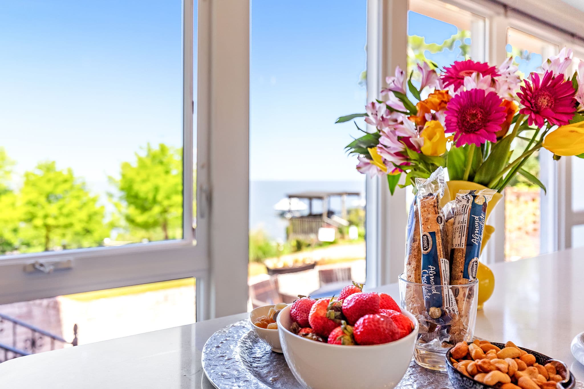A kitchen counter with fruit, snacks, and a vase of flowers, with a view of the ocean through large windows.