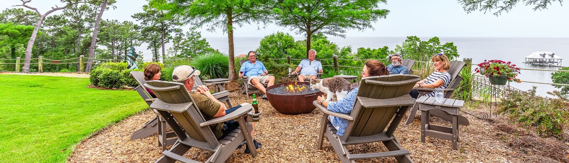 A group of people sitting in adirondack chairs around a fireplace in adirondack chairs visiting with a bay of water beyond them.