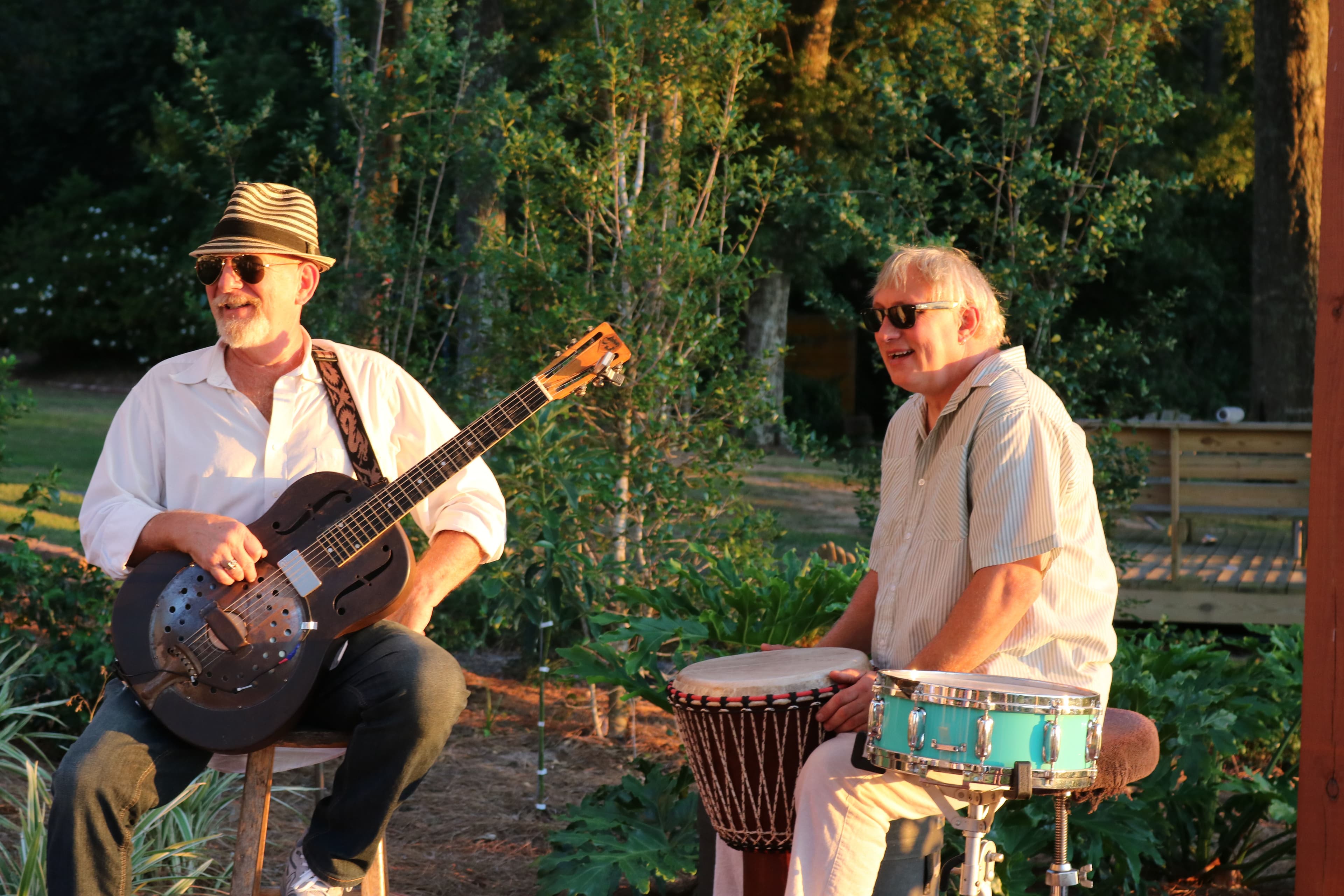 Two musicians play guitar and drums in a sunlit outdoor setting.