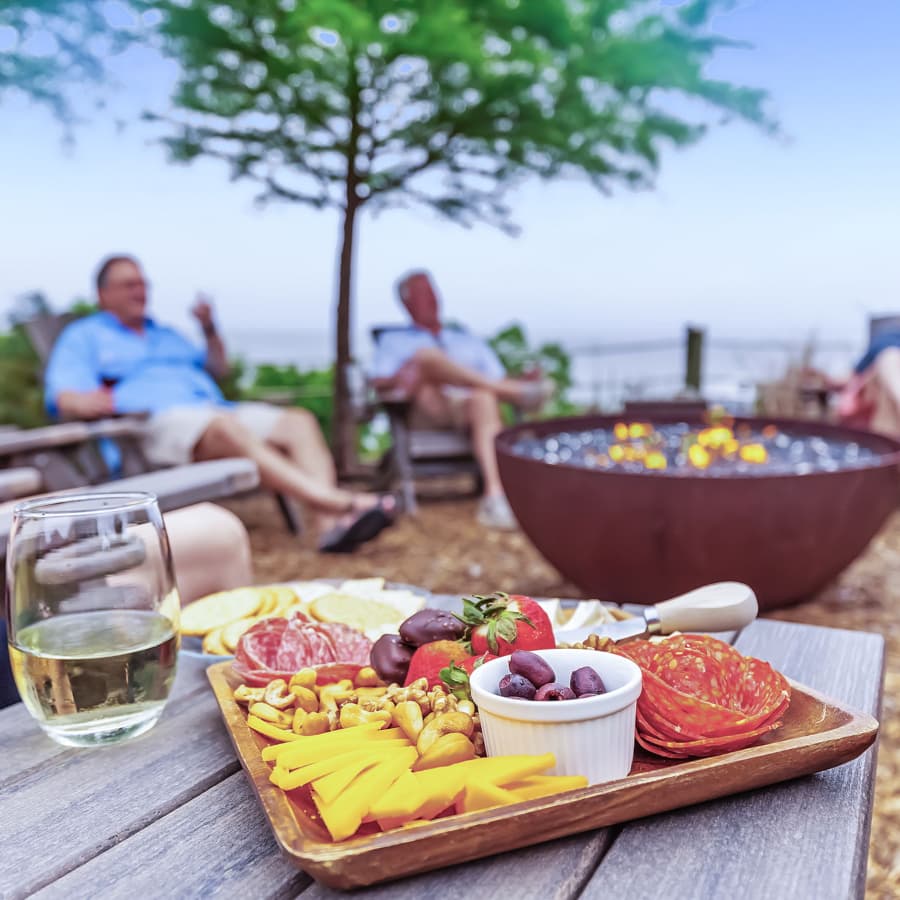 A charcuterie board filled with various meats, cheeses, olives and chocolates, along with a glass of white wine nearby, and a firepit in the background with people gathered around.
