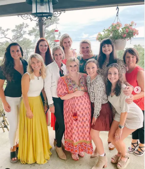 A group of twelve women poses together outdoors with plants and a scenic backdrop.