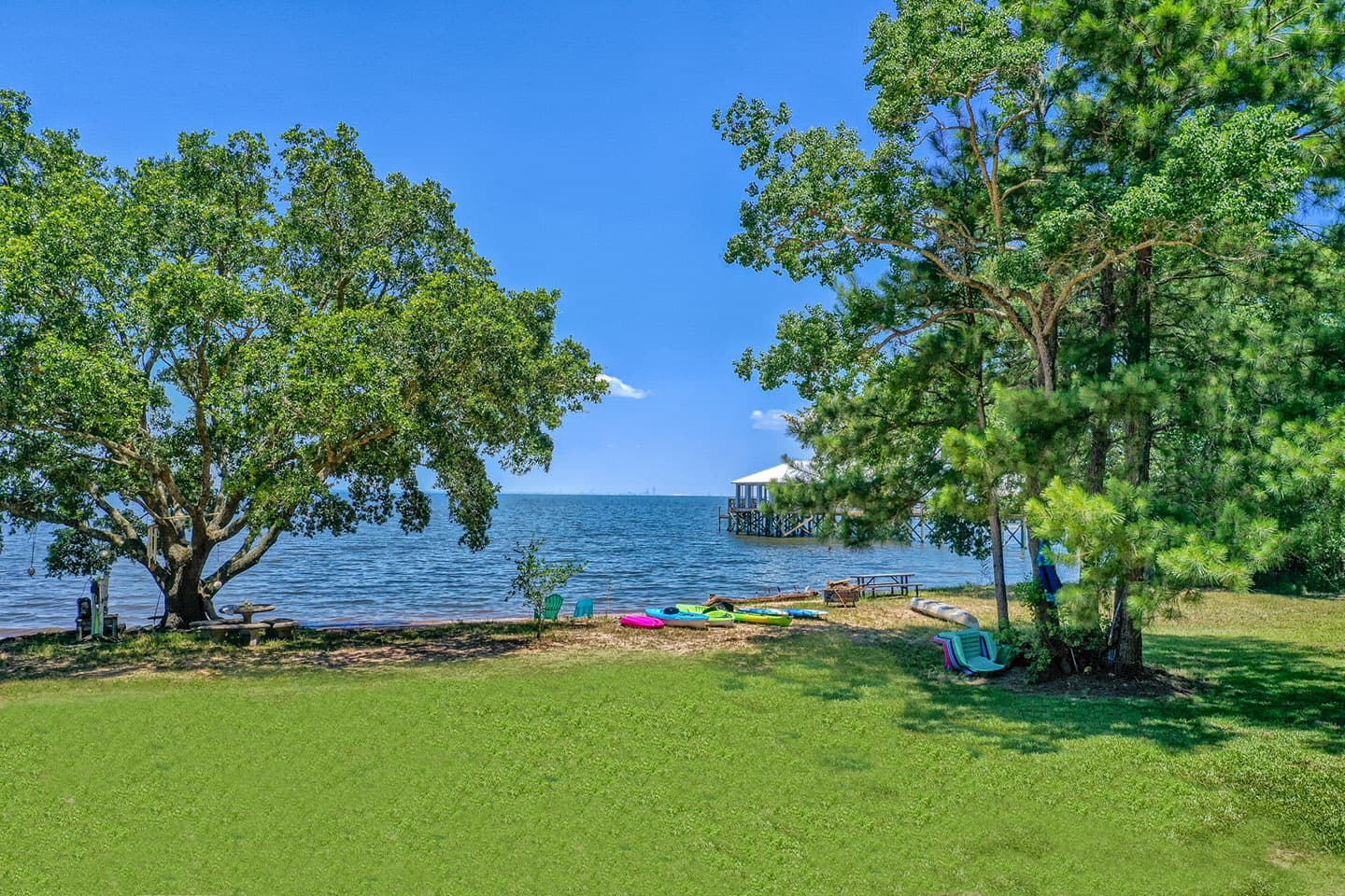 A picturesque bayside beach scene with lush green grass leading down to the water's edge. Tall trees frame the view, providing shade and creating a sense of seclusion. The sky is a brilliant blue with fluffy white clouds.
