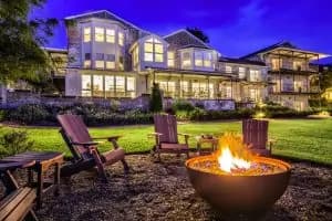 A cozy outdoor seating area with adirondack chairs surrounds a fire pit, with a modern house illuminated in the background under a twilight sky.