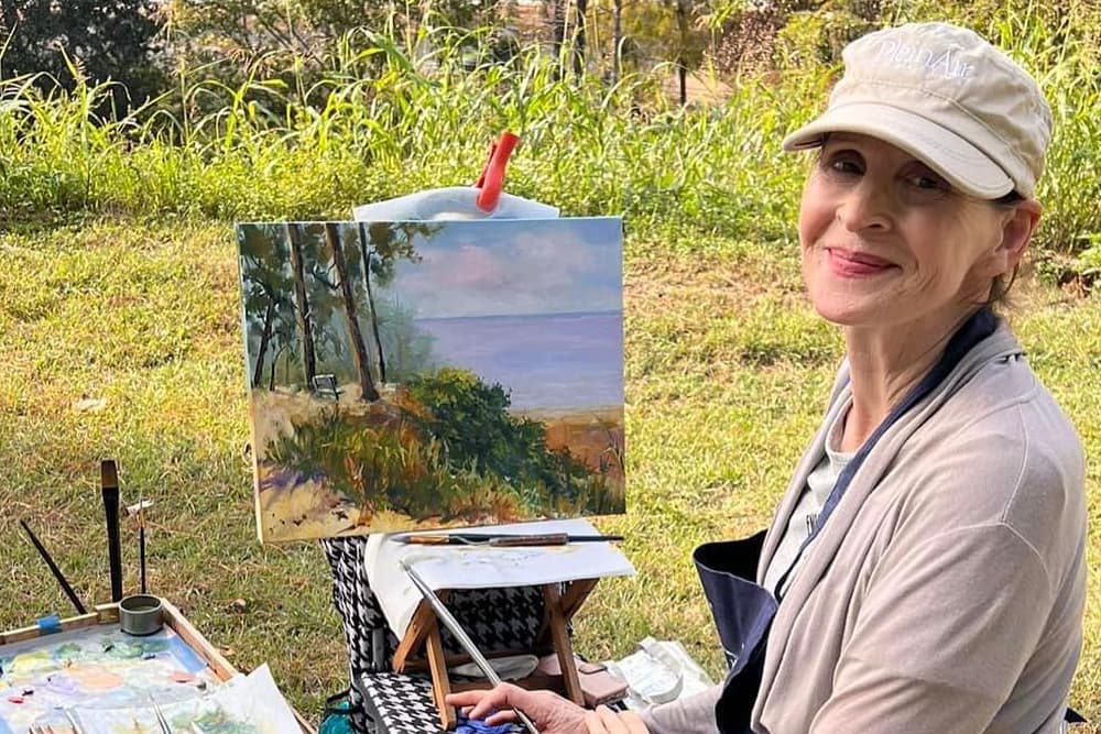 A smiling woman wearing an apron and hat painting a waterfront scene on canvas.