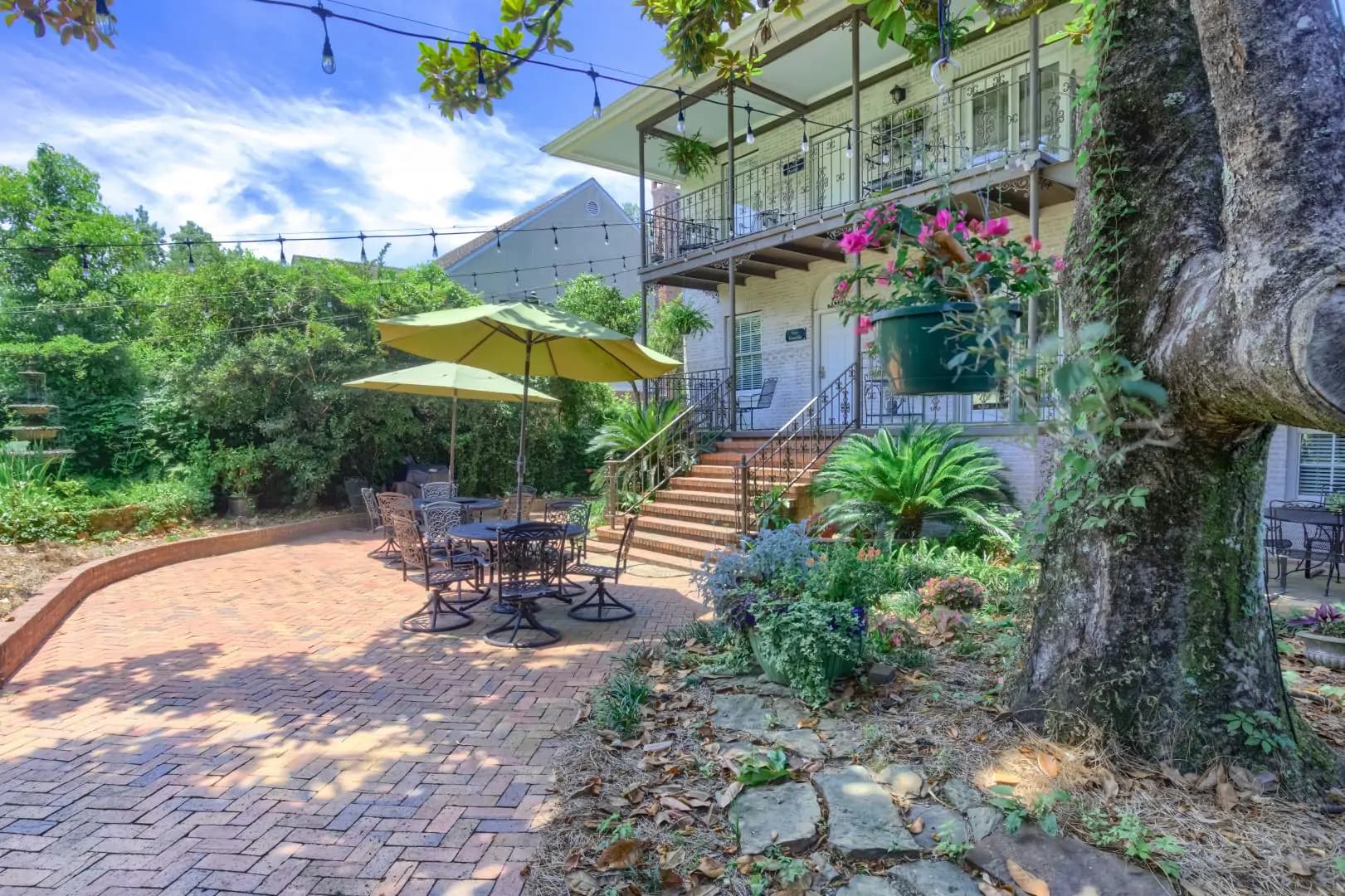A charming outdoor patio with a brick walkway, tables, and a large umbrella, surrounded by lush greenery and two-story building.