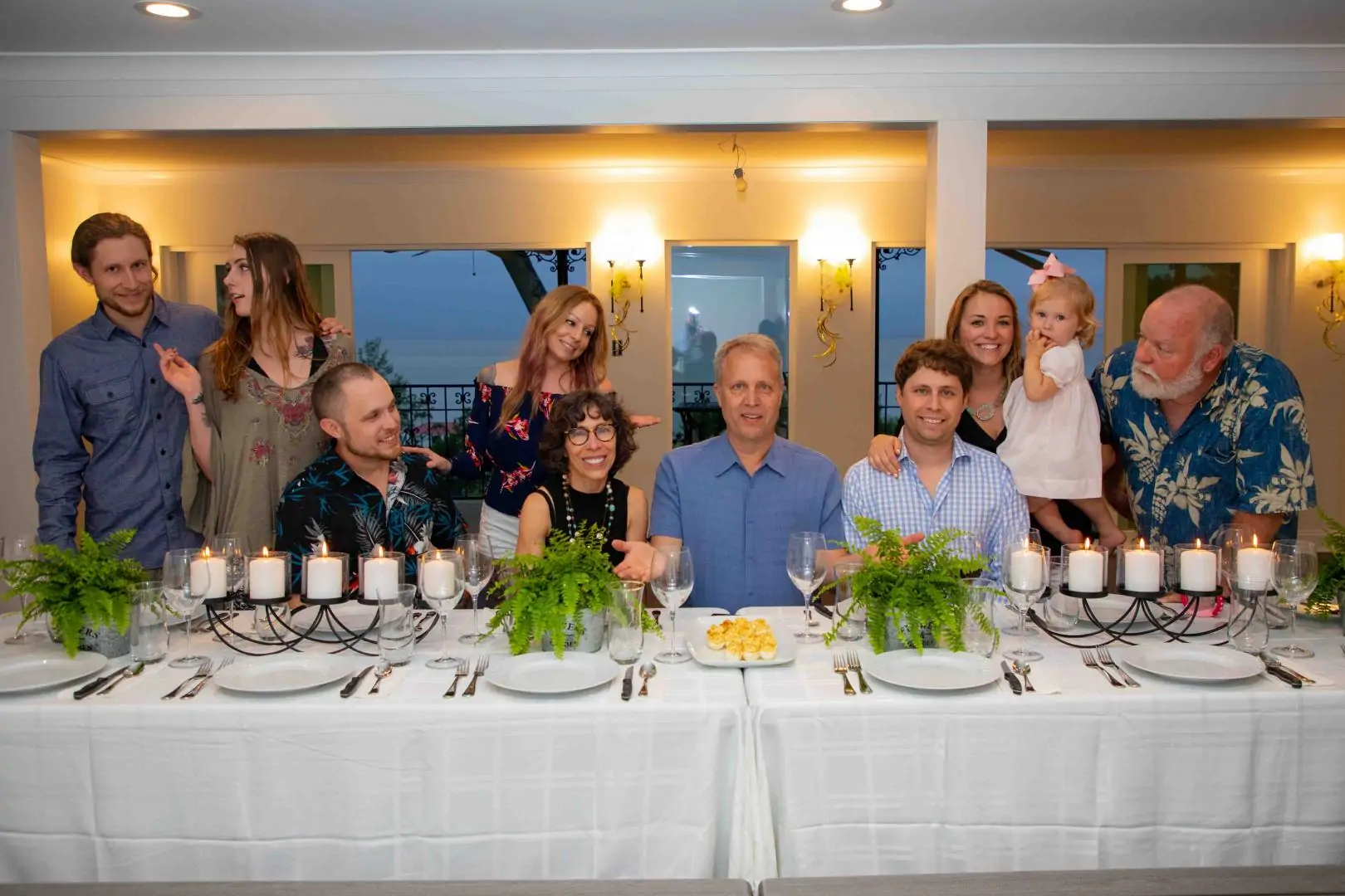 A group of seven adults and a child gather around a beautifully set dinner table, smiling and enjoying each other's company. A group of seven adults and a child gather around a beautifully set dinner table, smiling and enjoying each other's company.