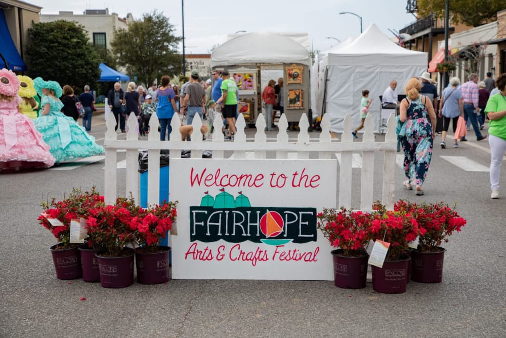 A street lined with vendors on either side and people strolling through the street perusing the vendors, and a white picket fence in front with a sign in front of the fence that says Welcome to the Fairhope Arts & Crafts festival with pots of red flowers on either side.