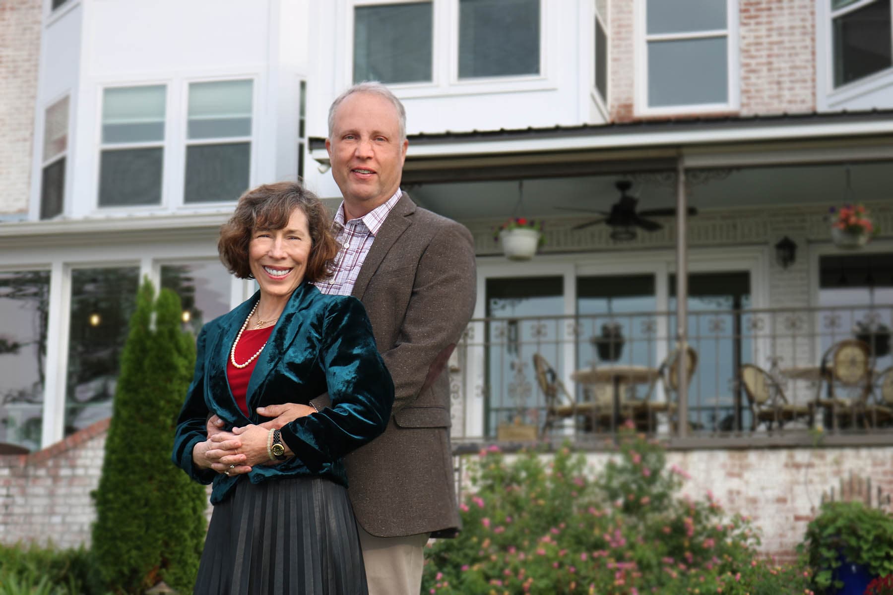 A smiling couple stands together in front of a quaint house with a porch. A smiling couple stands together in front of a quaint house with a porch.
