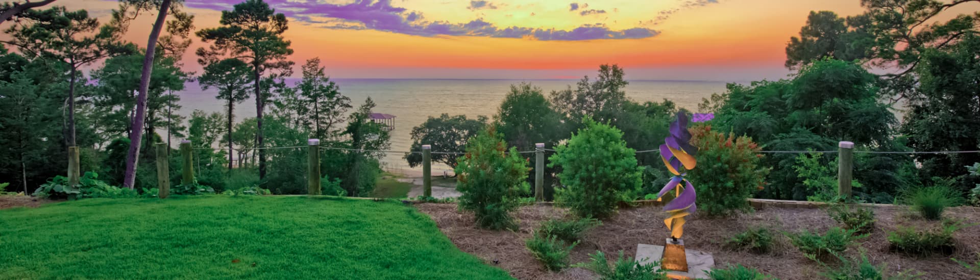 A vibrant sunset over water, framed by lush greenery and a colorful sculpture in the foreground.