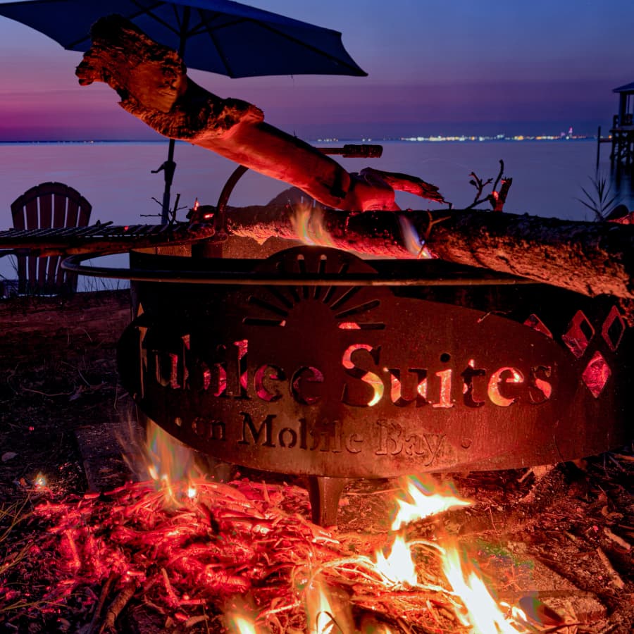 An evening fire in a Jubilee Suites metal fire ring with chairs nearby and the bay in the background