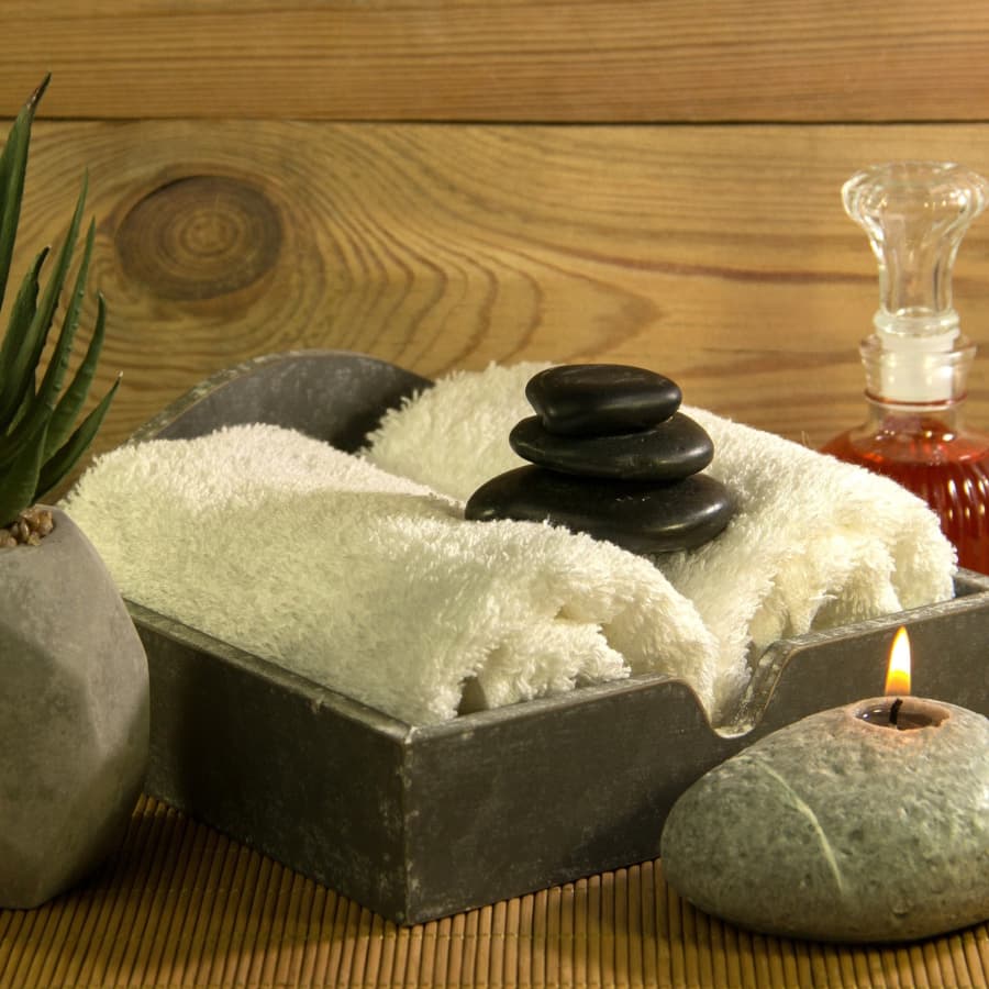 A wood tray filled with two rolled towels, a small stack of rocks on top of them, a rock with a tealight in front of them, a plant to one side and a bottle of massage oil on the other side.