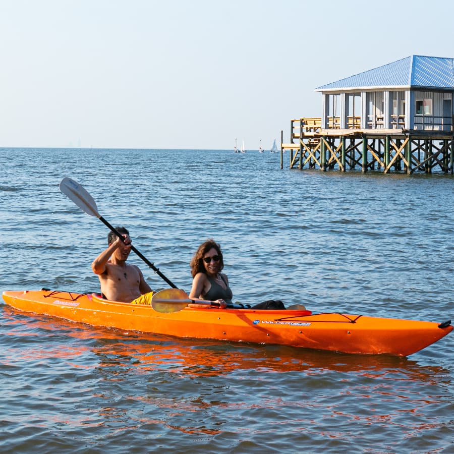 A man and woman in an orange kayak on the waters of Mobile Bay with a house on stilts n the water and several sailboats in the distance.