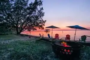 A peaceful lakeside scene at sunset featuring a fire pit, colorful chairs, and umbrellas.