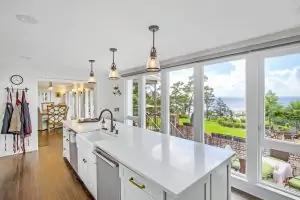 Bright, modern kitchen with a white island and large windows offering a view of greenery and the ocean.