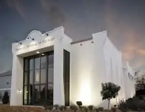 A modern white building with large glass windows and a decorative facade, set against a twilight sky.