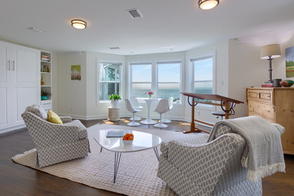 A spacious living room are in a bedroom featuring a curved bay window with ocean views, comfortable seating, a dining table and 2 chairs, a dresser, and an artists drawing board with stool.