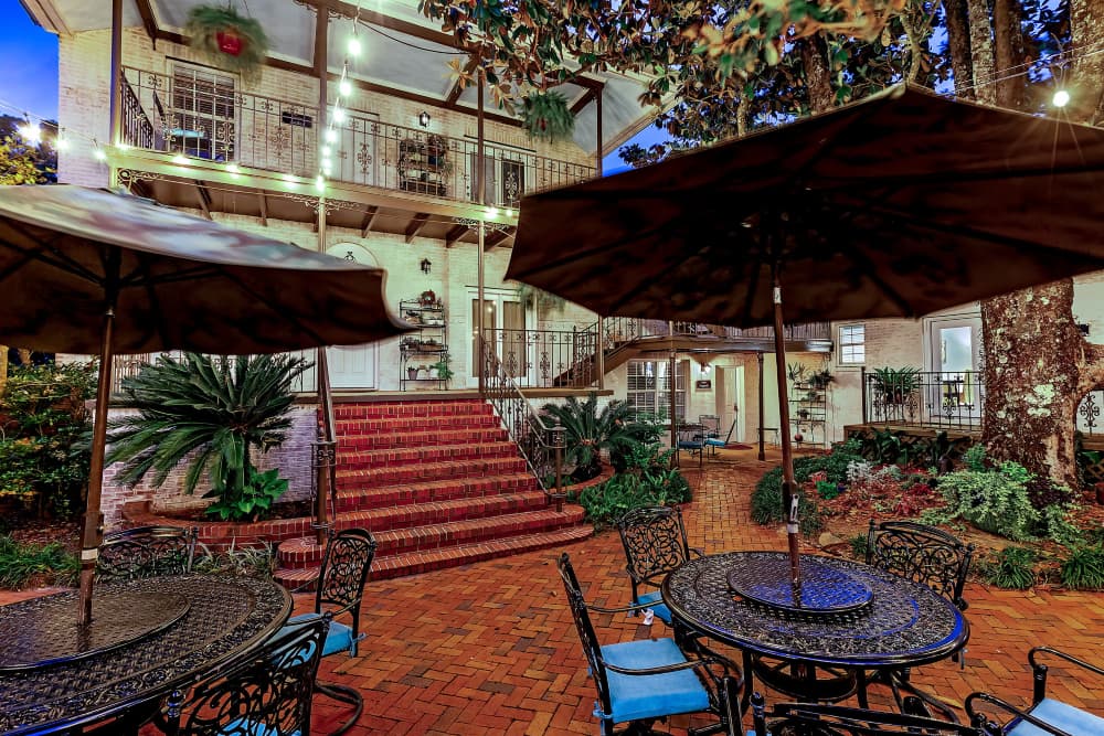 A welcoming courtyard with red brick floors, two wrought iron tables and chairs with blue cushions and blue sun umbrellas, a large shade tree and numerous bushes and plants, and multi-story houses on 2 sides of the courtyard, reminiscent of the New Orleans style architecture.