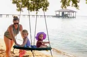 A woman pushes two children on a swing by the beach, with a pier and a gazebo in the background.
