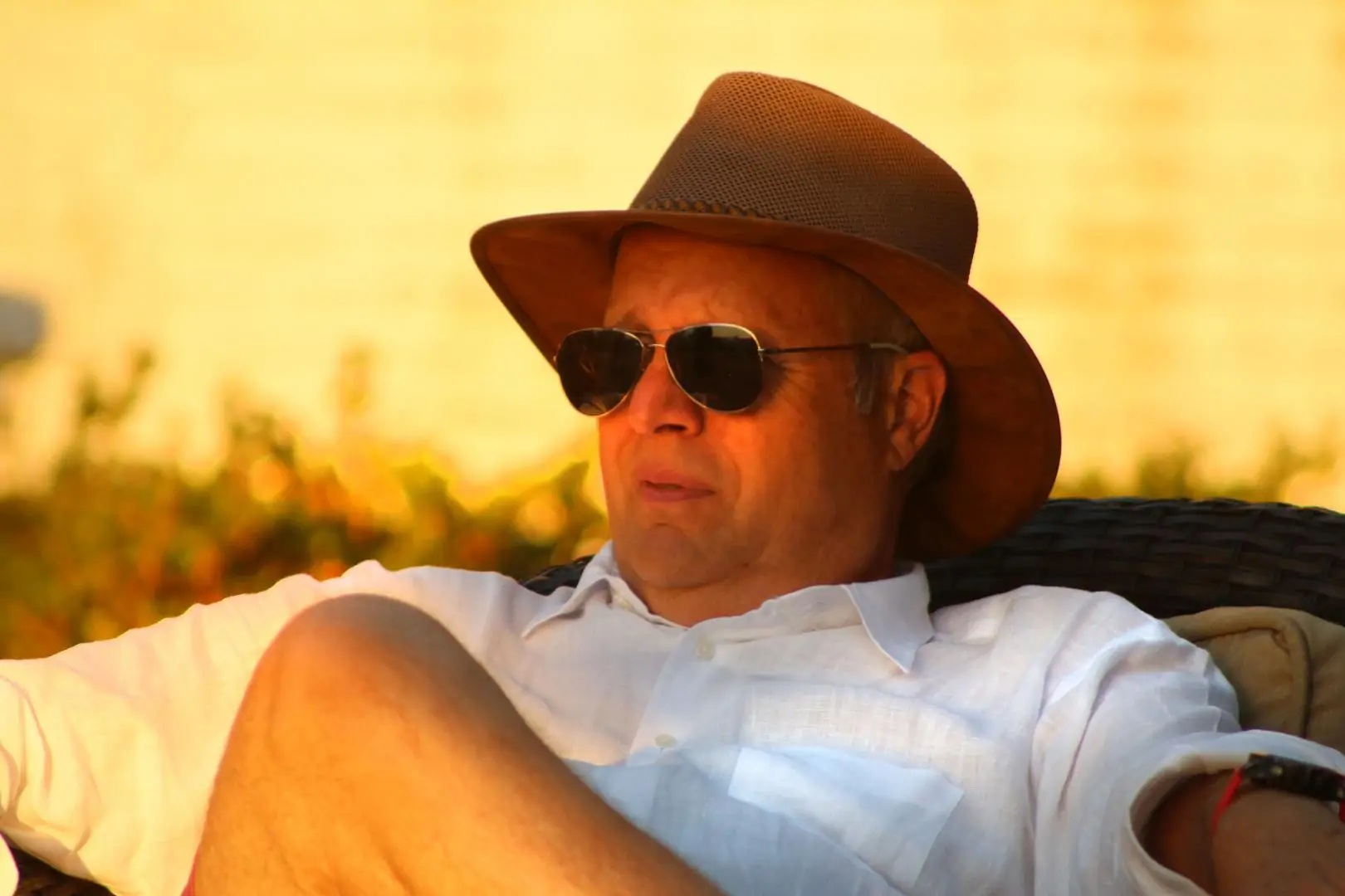 A man wearing sunglasses and a hat relaxes outdoors in warm sunlight.
