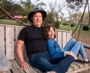 A man and woman sit together on a wooden swing in a park.