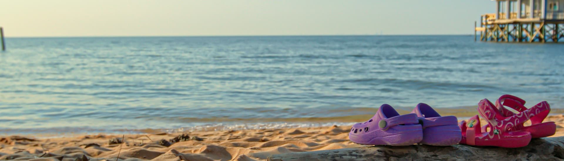 A sandy beach with 2 pairs of colorful crocs on the sand, with the waters of Mobile Bay inviting you to come explore, and a pier on one side.