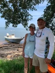 A couple stands by a boat near the shore on a sunny day.