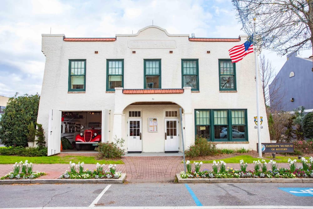 A white stucco building with a red tiled roof with a vintage car in the garage and a sign out front that says Fairhope Museum of History