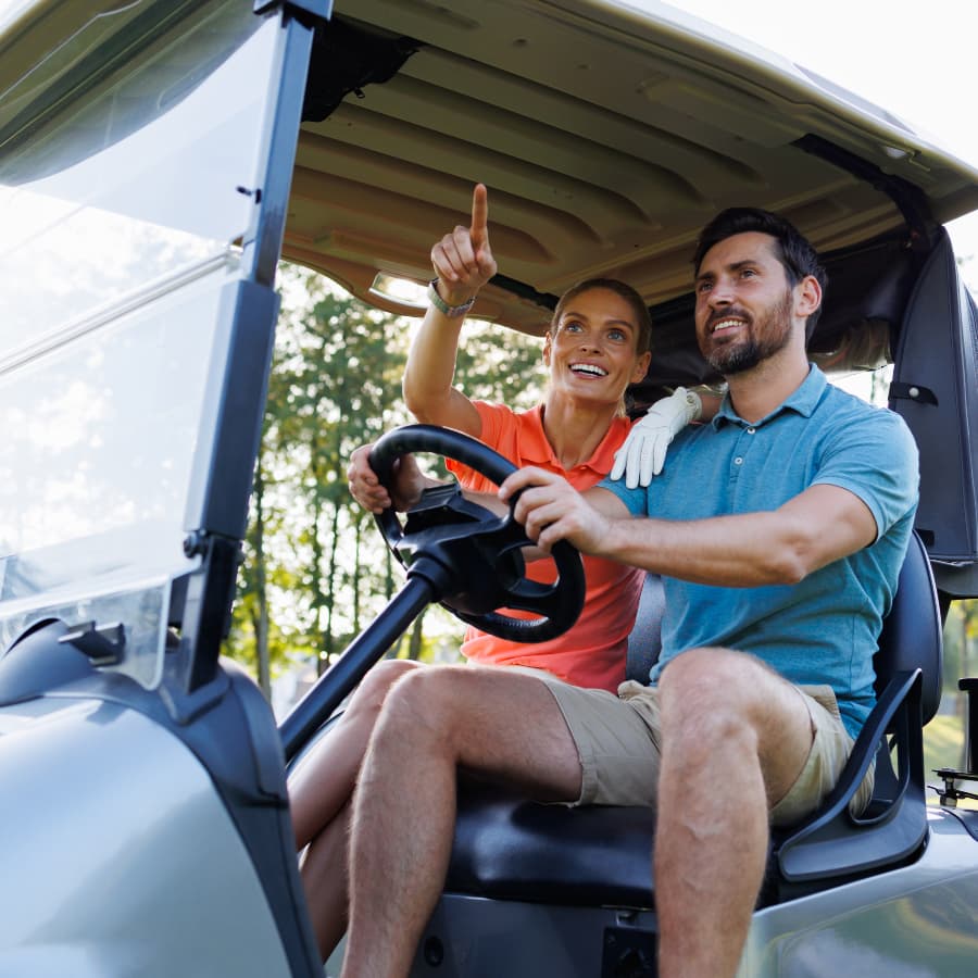 A man and a woman in a golf cart, the man driving the cart, and the woman pointing at something in the distance.