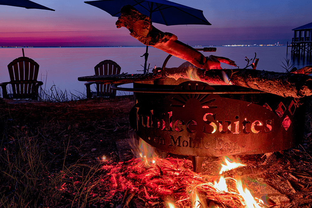 Campfire glowing by the water's edge under a twilight sky, featuring chairs and an umbrella nearby.