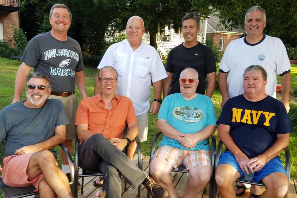 A group of eight men of varying ages smiles for a photo outdoors, seated in two rows on a wooden deck.