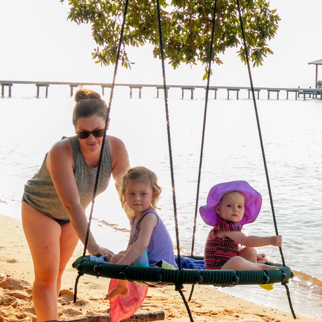 A mother pushing her two young children in a round swing in front of Mobile Bay.